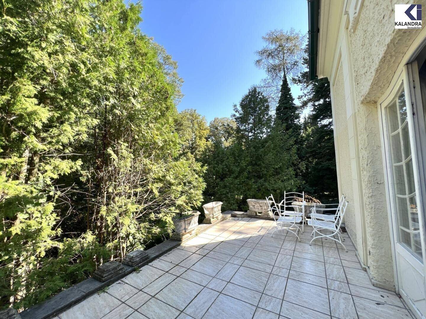 Sonniger Terrassenbereich mit Fliesenboden und Gartenmöbeln, umgeben von üppigem Grün.
