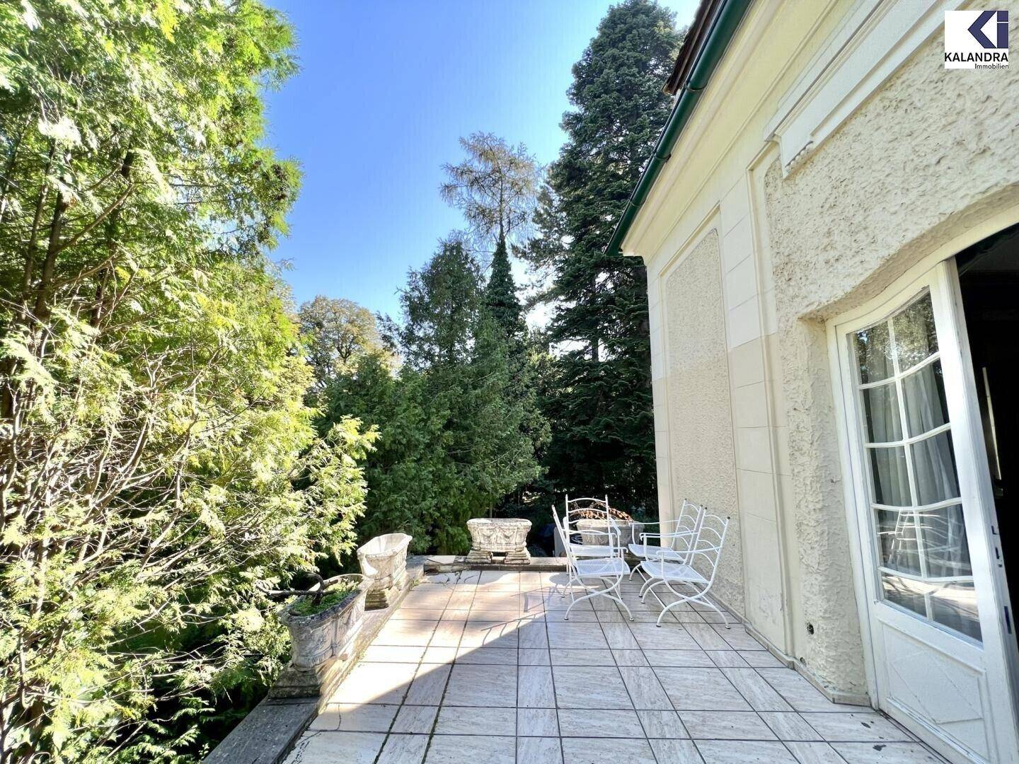 Helle Terrasse mit Fliesenboden und Sitzgelegenheiten, umgeben von hohen Bäumen und Sträuchern.