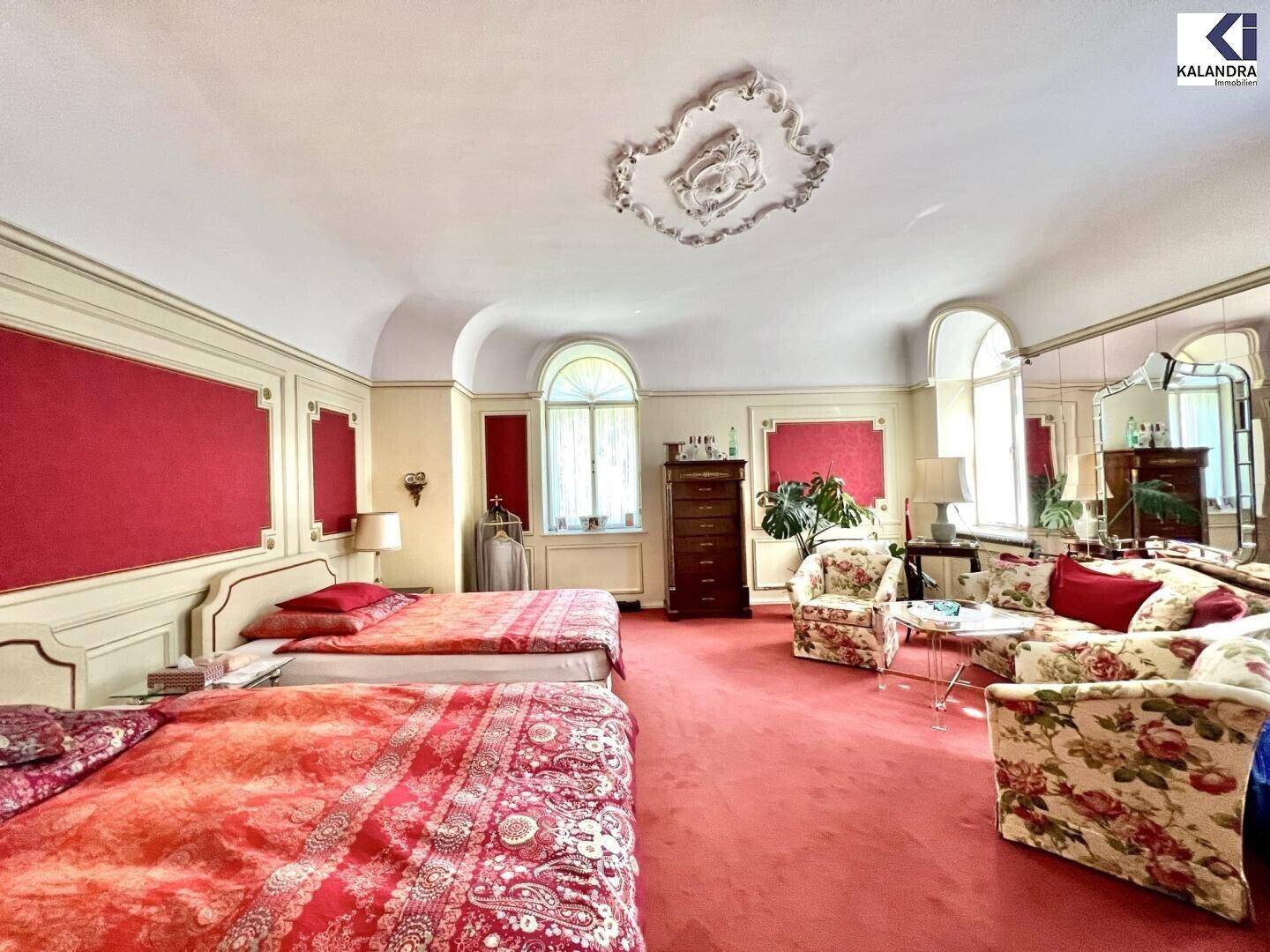 Geräumiges Schlafzimmer mit roten Akzenten, Stuckdecke und zwei großen Betten.