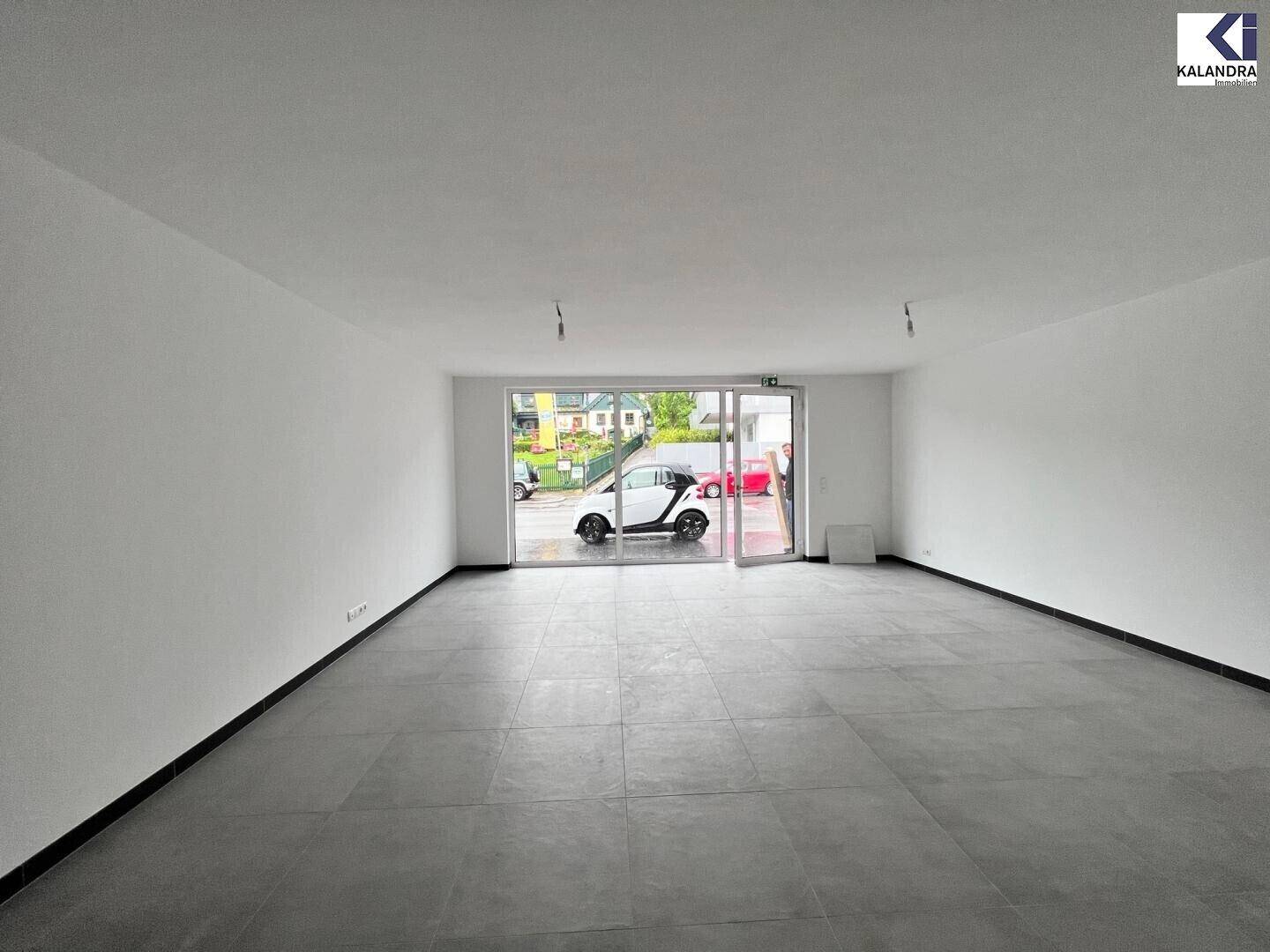 Heller, leerer Raum mit großen Glasfronten zur Straße hin, durch die ein weißes Auto sichtbar ist.