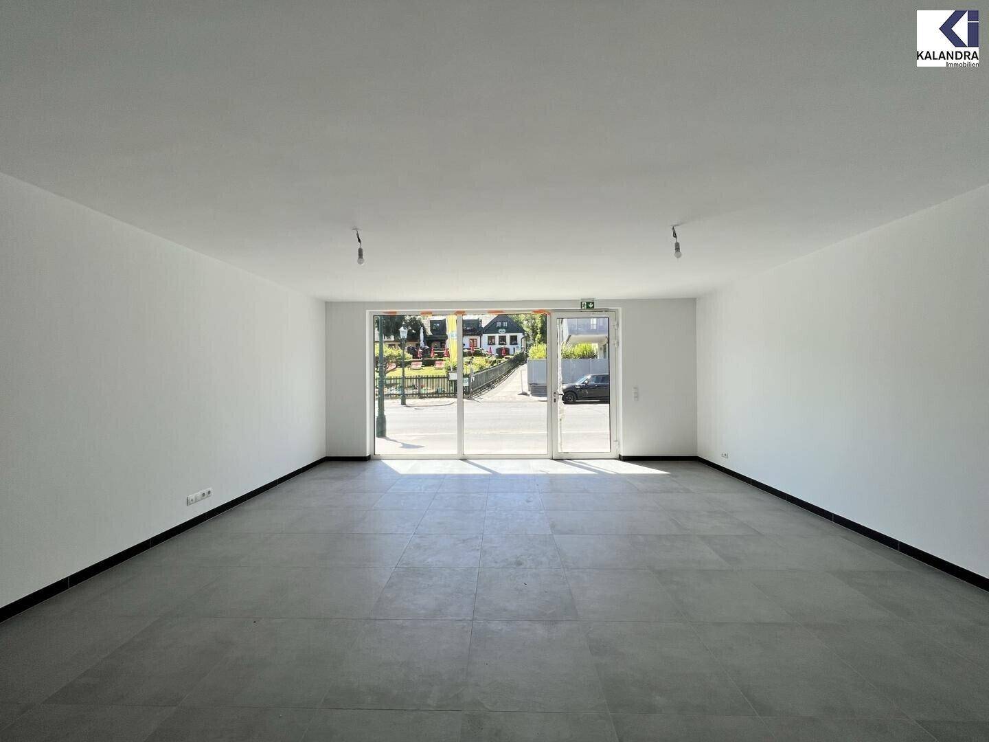 Heller, leerer Raum mit großen Glasfronten zur Straße hin und modernem Fliesenboden.