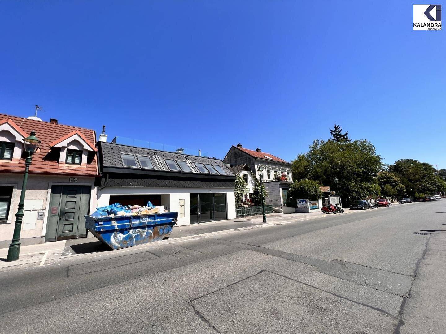 Weitläufige Straßenansicht mit dem modernen Gebäude, angrenzenden Häusern und einem blauen Container.