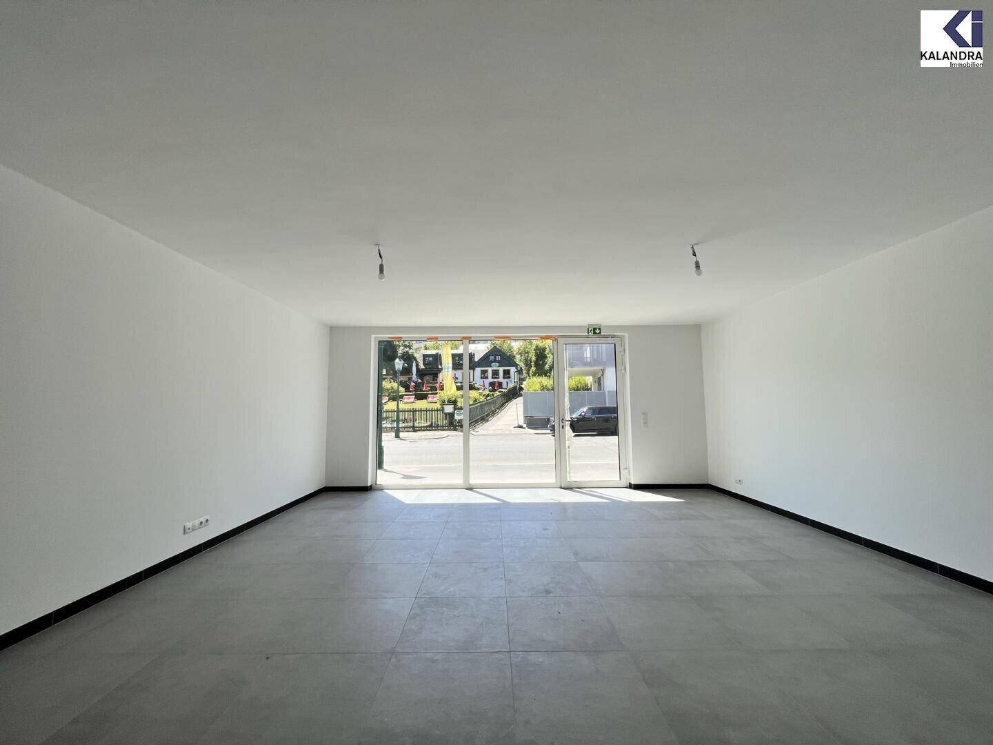 Heller, leerer Raum mit großen Glasfronten zur Straße hin und modernem Fliesenboden.