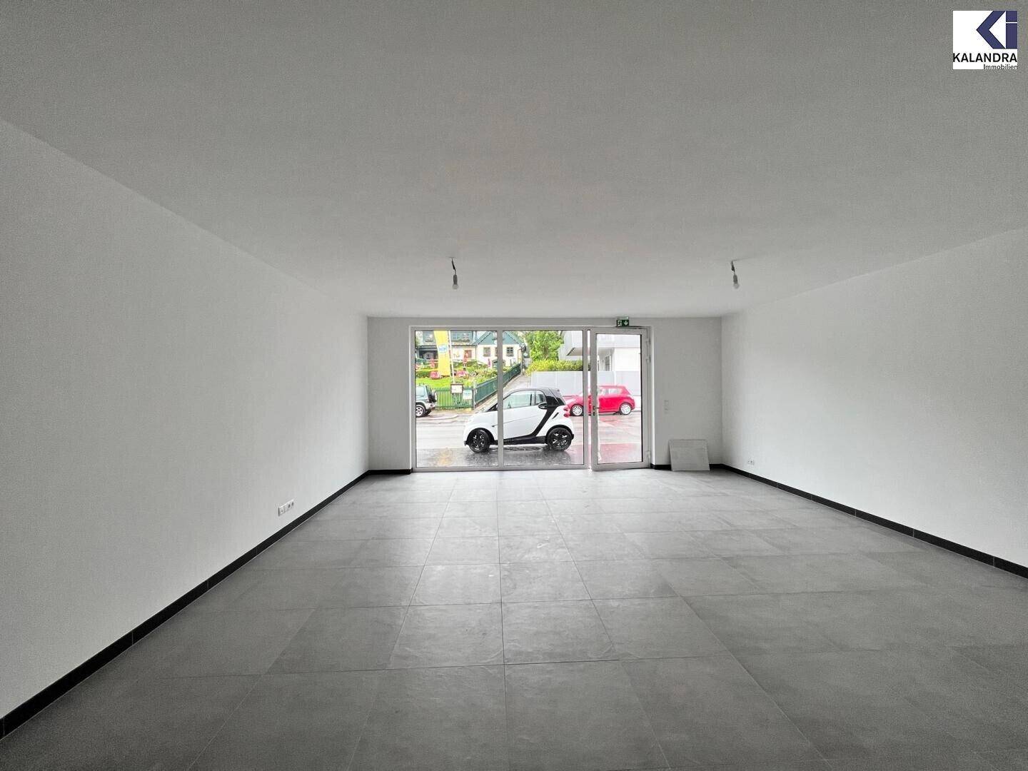 Heller, leerer Raum mit großen Glasfronten zur Straße hin, durch die ein weißes Auto sichtbar ist.