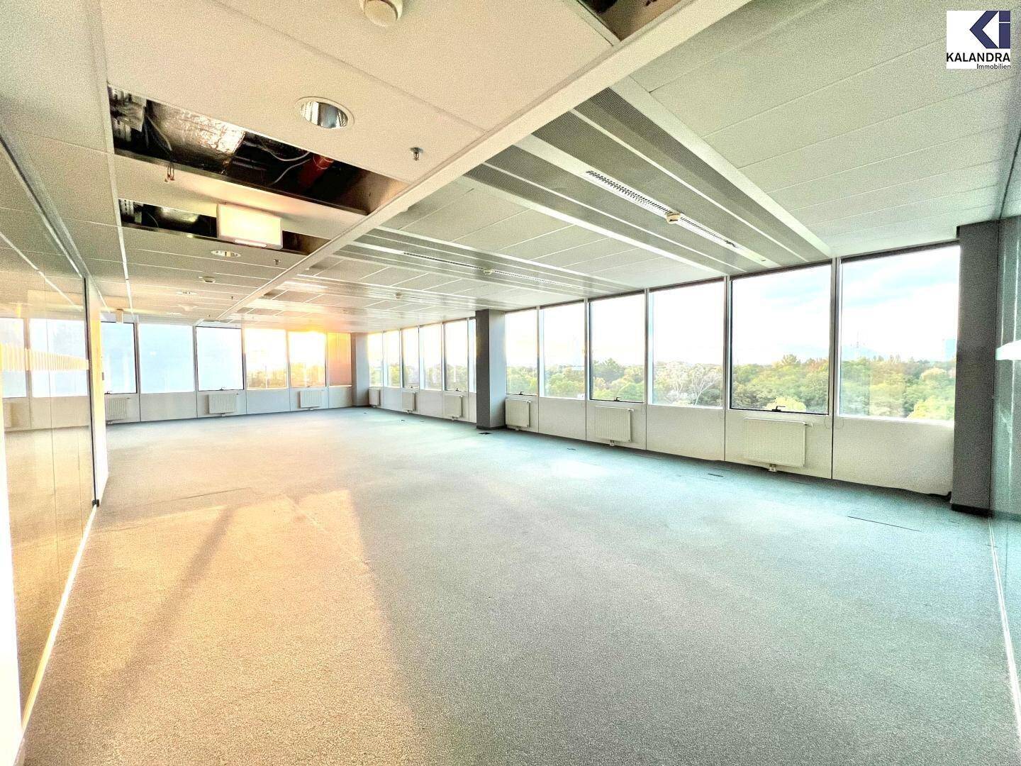Helles, leeres Büro mit Teppichboden und großen Fenstern, die von der Sonne durchflutet werden.