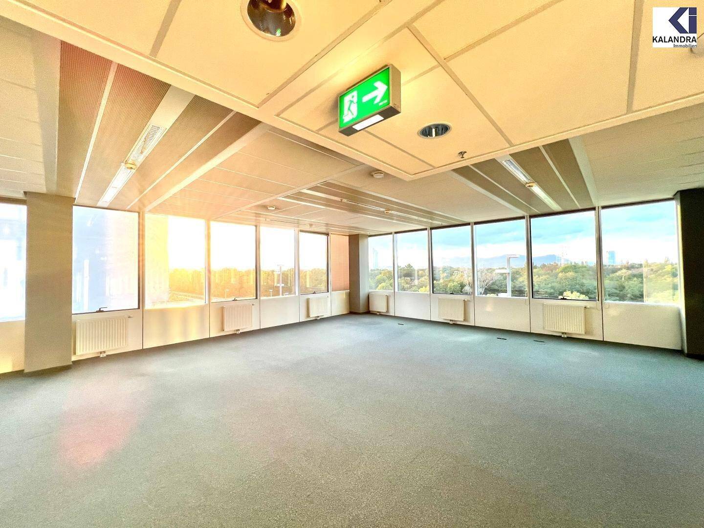 Helles, leeres Büro mit Teppichboden und großen Fenstern, die von der Sonne durchflutet werden.