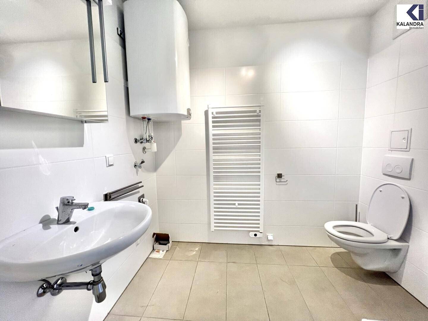 Helles Badezimmer mit weißen Fliesen, Waschbecken, Toilette und Handtuchheizkörper.