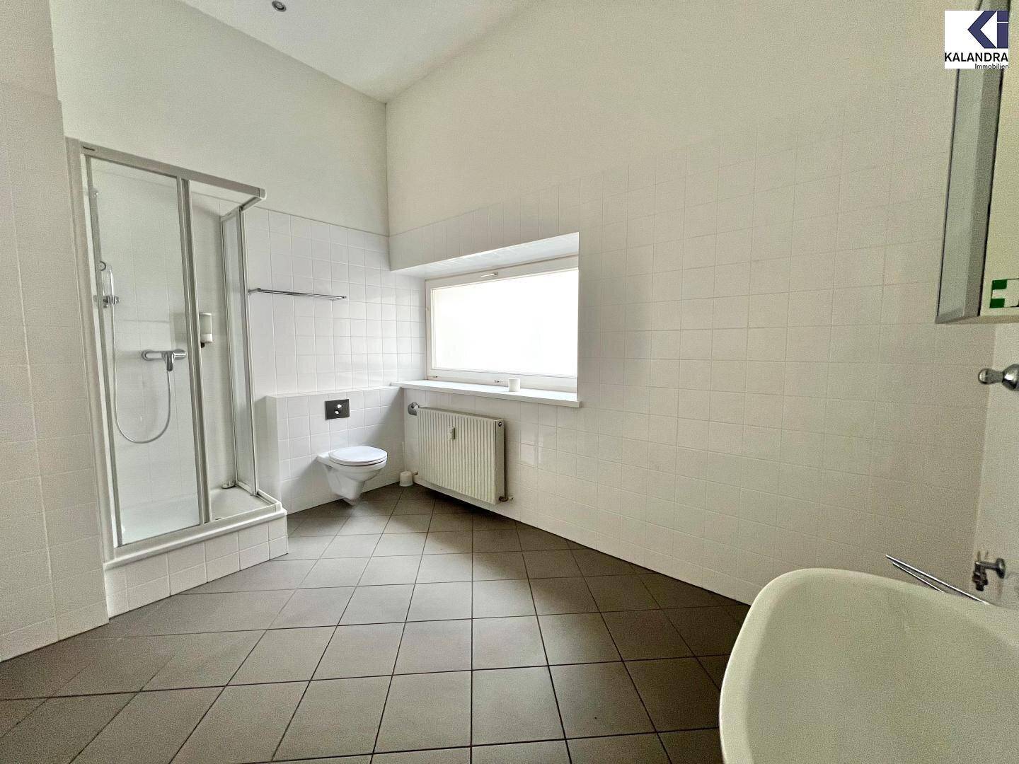 Badezimmer mit Duschkabine, WC, Waschbecken und Fenster, komplett gefliest.