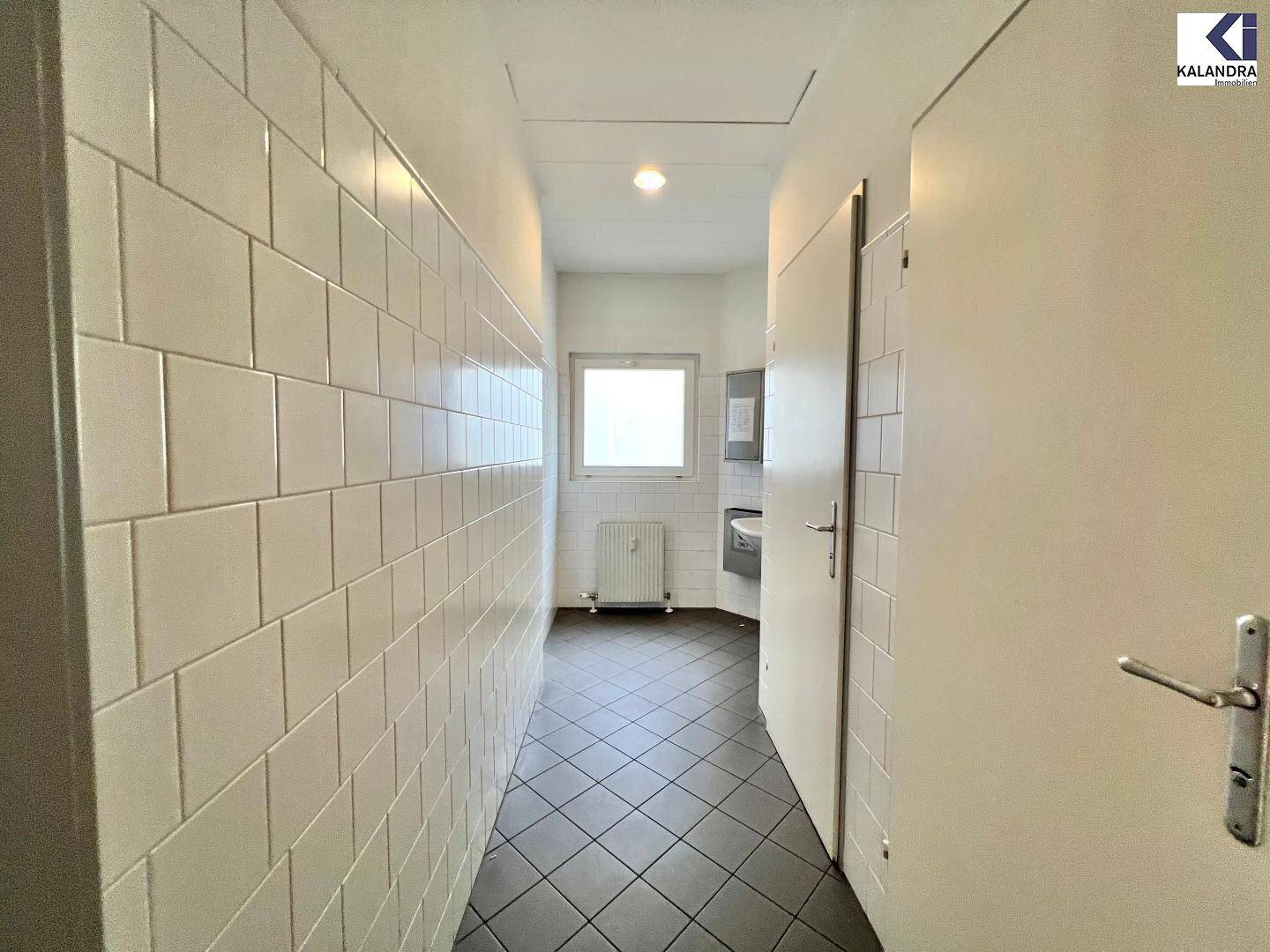 Gefliester Flur mit weißen Wandfliesen, der zu einem Badezimmer mit Fenster führt.