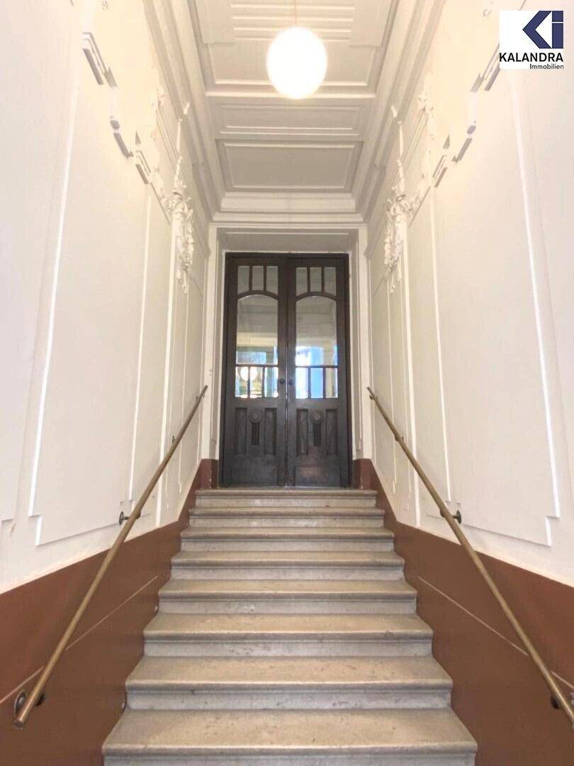 Repräsentativer Eingangsbereich mit breiter Steintreppe, verzierten Geländern und einer hohen Decke mit Stuckelementen.