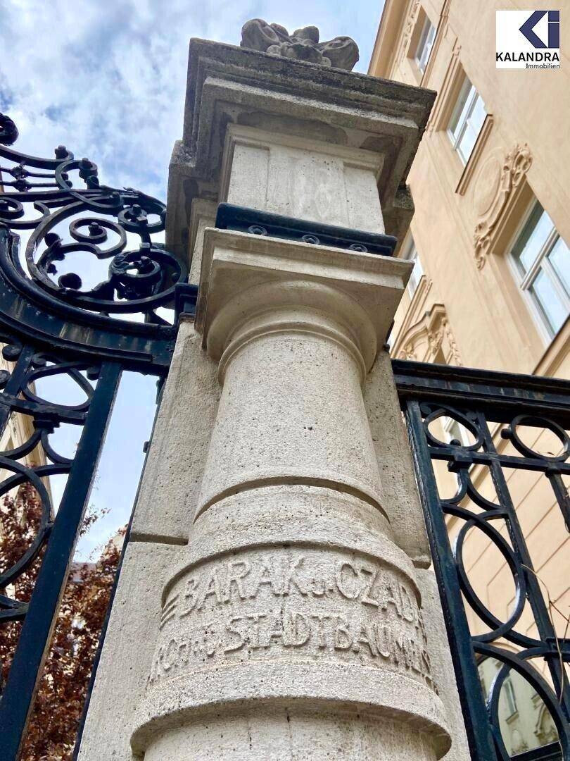 Detailansicht eines historischen Steinsockels mit Inschrift und schmiedeeisernem Zaun vor einem Altbau.
