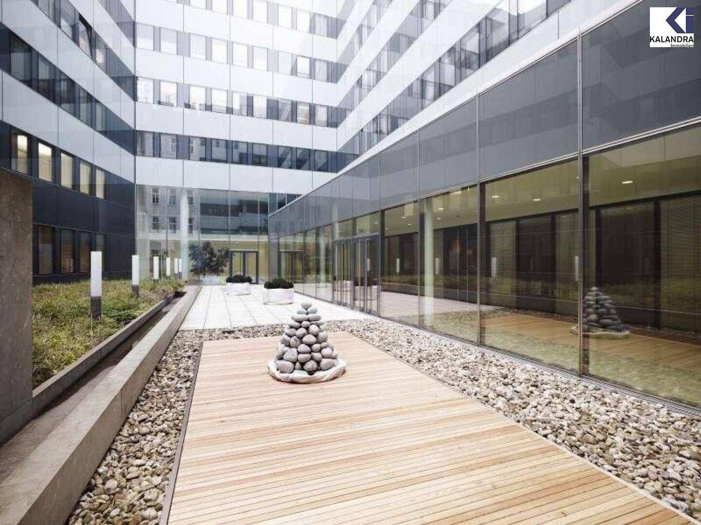 Moderner Innenhof mit Holzterrasse, Kiesflächen, Grünbepflanzung und reflektierenden Glasfassaden.