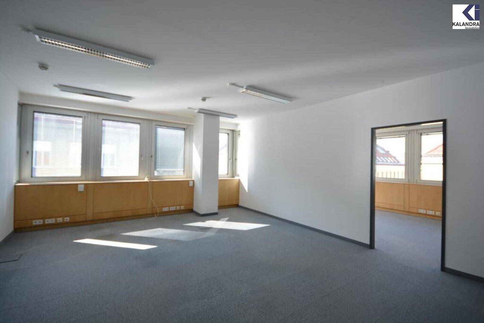 Heller Büroraum mit Teppichboden, Fenstern mit Jalousien und einem Durchgang zu einem weiteren Raum.