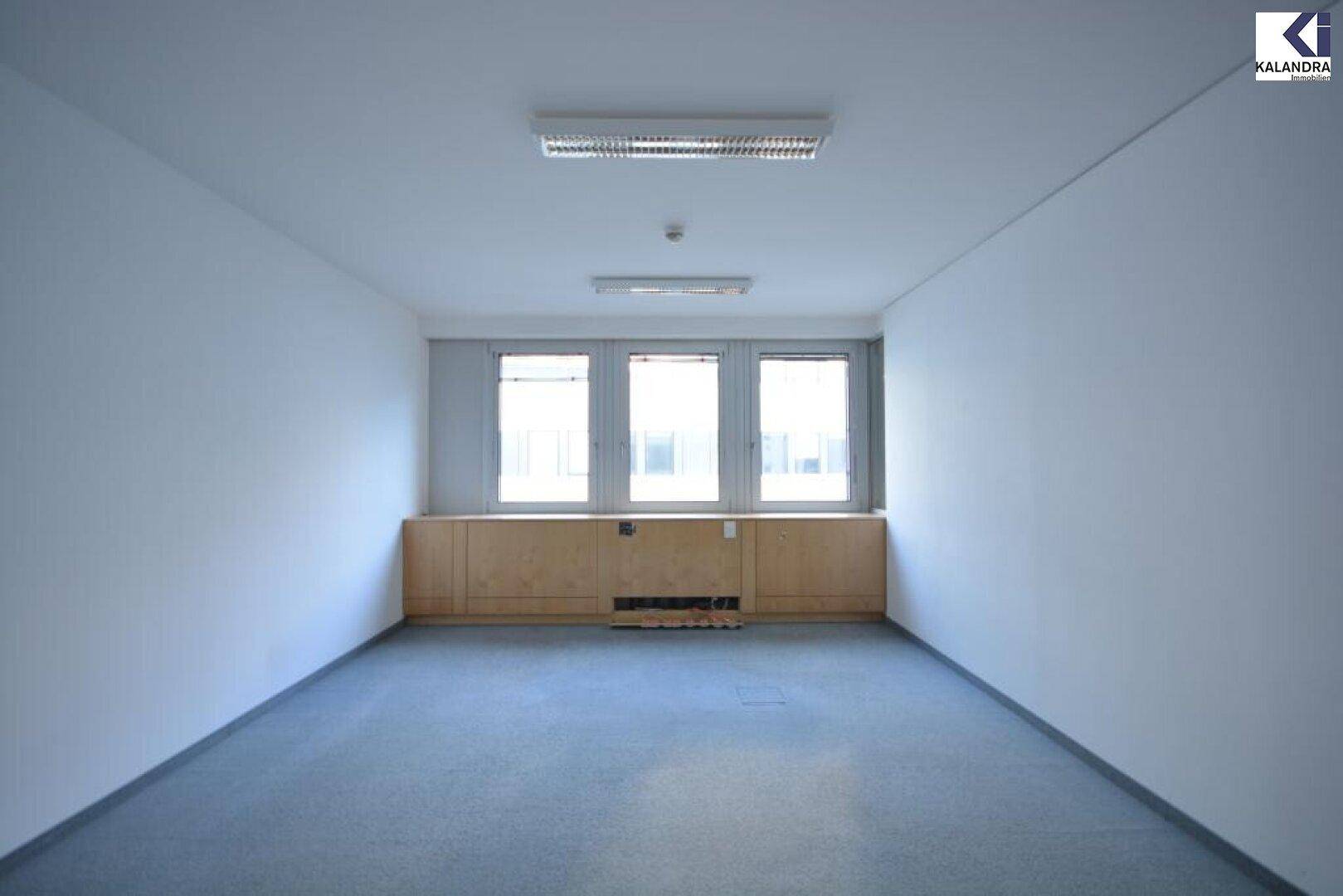Langer, schmaler Büroraum mit Teppichboden, weißen Wänden und drei Fenstern am Ende des Raumes.