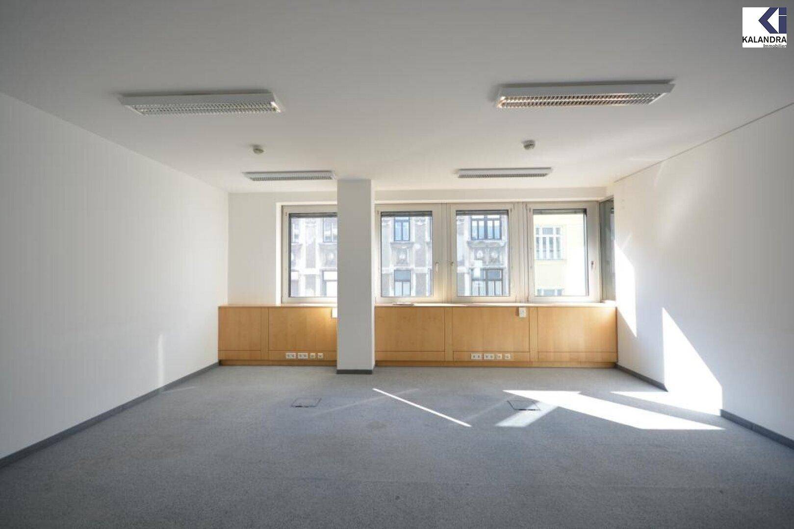 Heller Büroraum mit Teppichboden, großen Fenstern und Holzverkleidung unter den Fenstern.