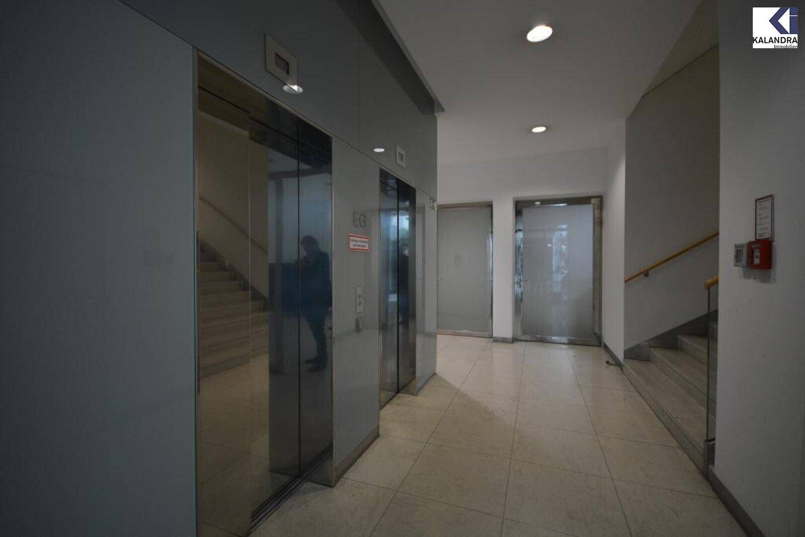 Moderner Flur mit zwei Aufzügen, Treppenhaus und Fliesenboden in einem Bürogebäude.