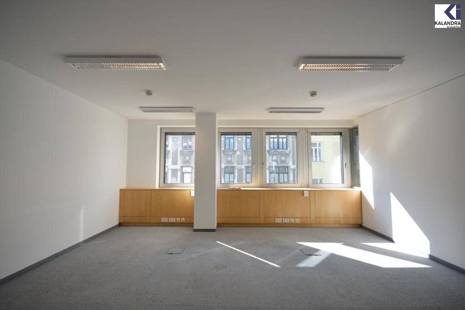 Heller Büroraum mit Teppichboden, großen Fenstern und Holzverkleidung unter den Fenstern.