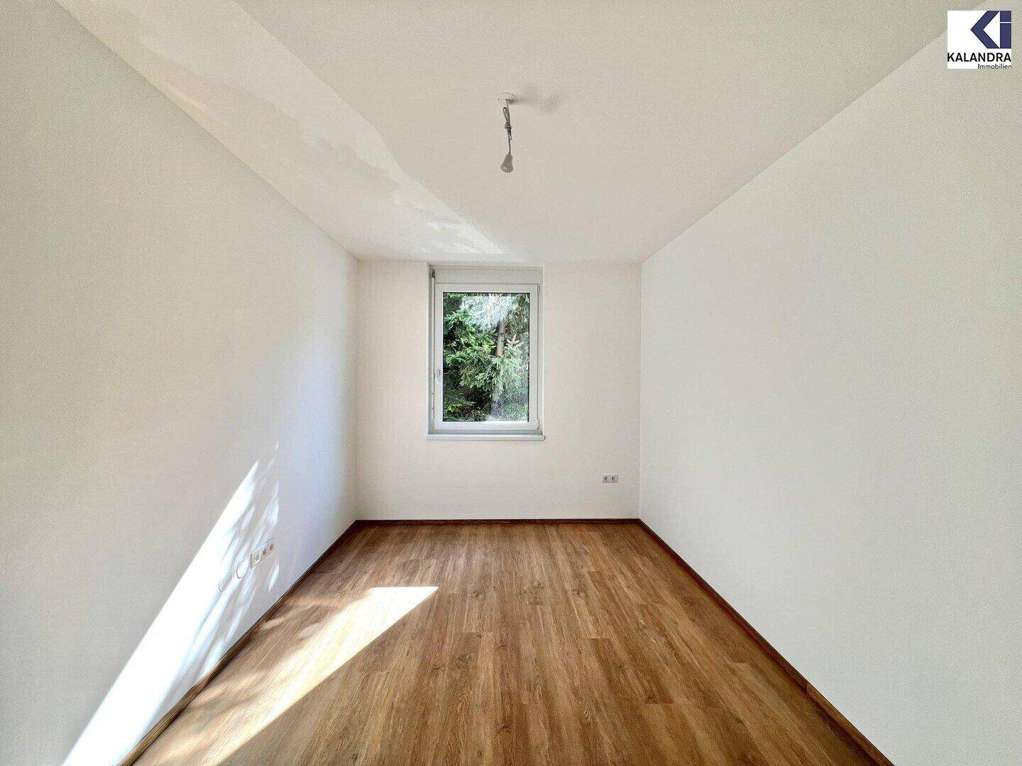 Leeres Zimmer mit hellem Holzboden, weißen Wänden und einem Fenster mit Blick ins Grüne.