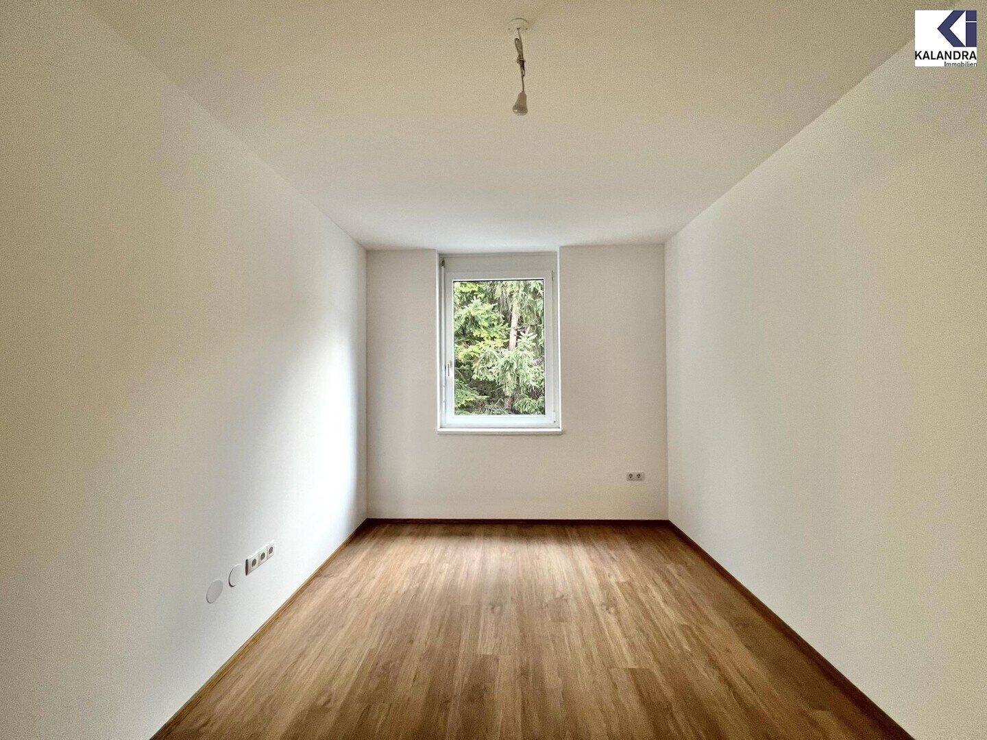 Leeres Schlafzimmer mit Holzboden, weißen Wänden und einem Fenster mit Blick ins Grüne.