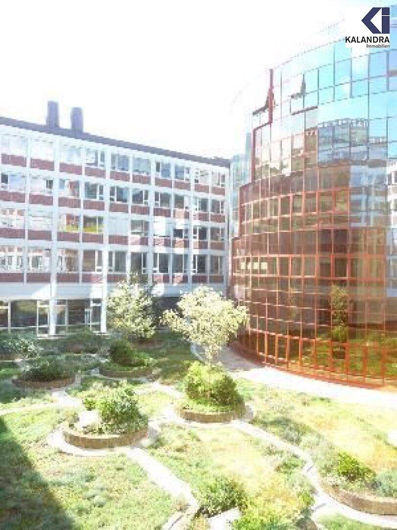 Innenhof mit Grünflächen, umgeben von einem modernen Gebäude mit Glasfassade und einem Bürogebäude.
