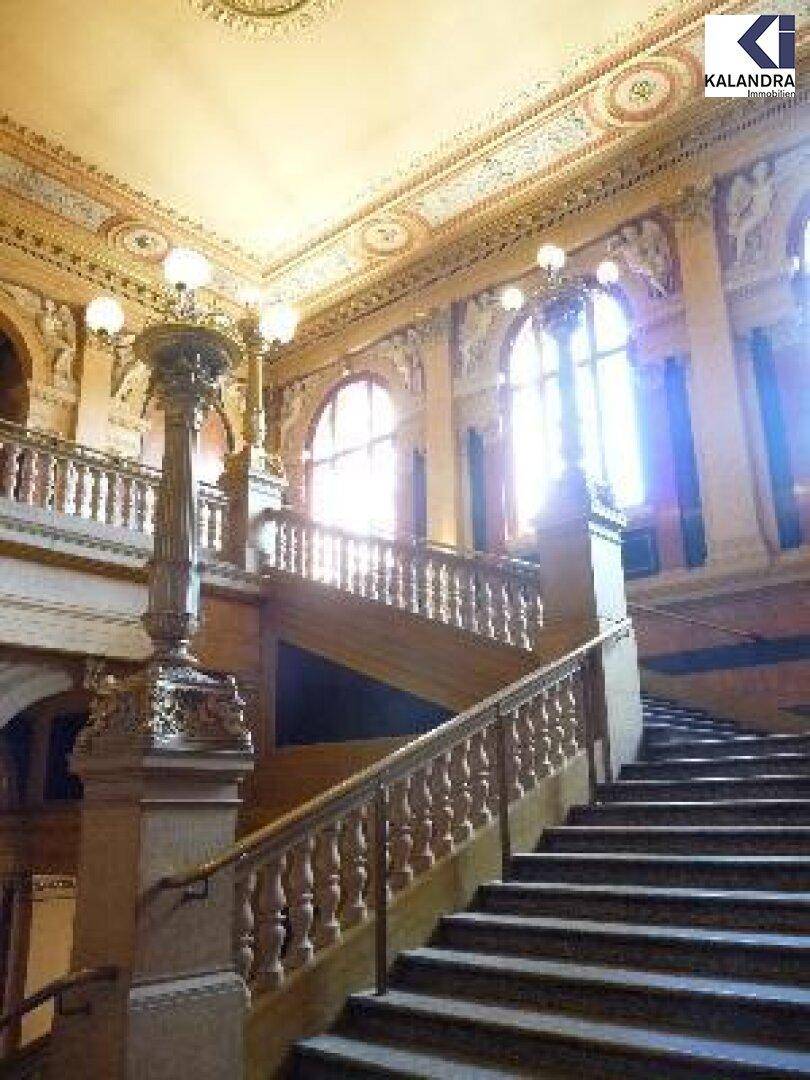 Eindrucksvolles Treppenhaus mit kunstvollen Verzierungen, hohen Decken und großen Fenstern.