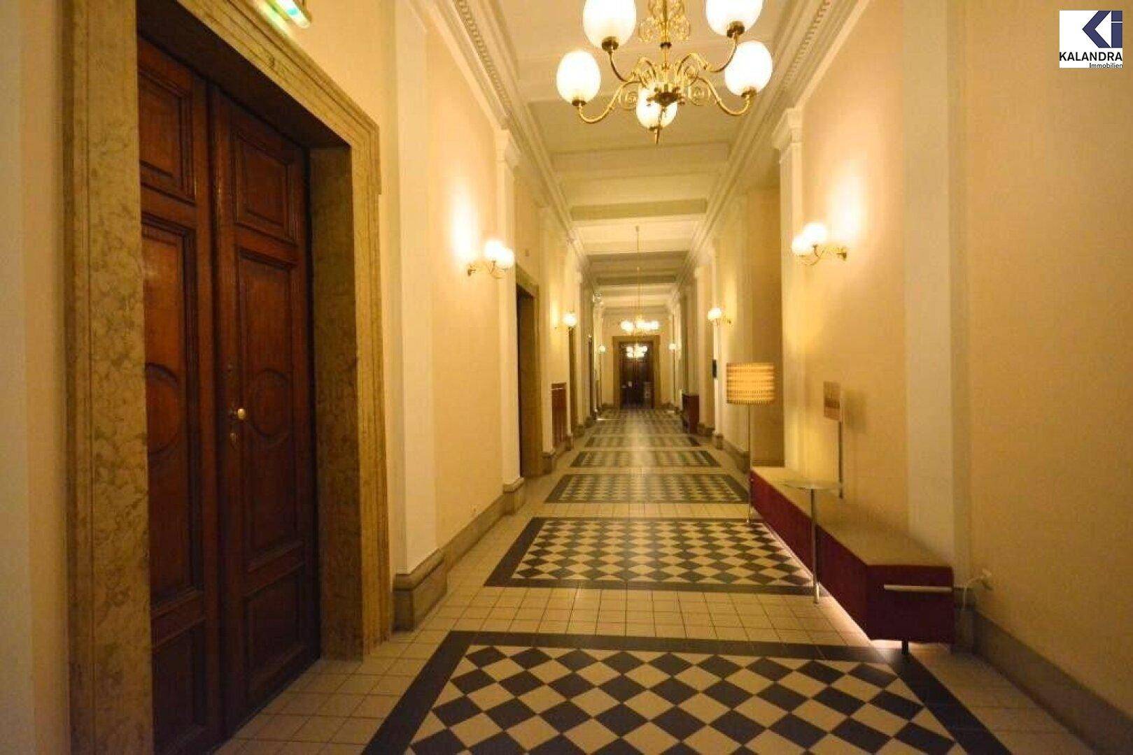 Eleganter Flur mit Schachbrettmuster-Fliesenboden, Kronleuchtern und Wandleuchten in einem historischen Gebäude.