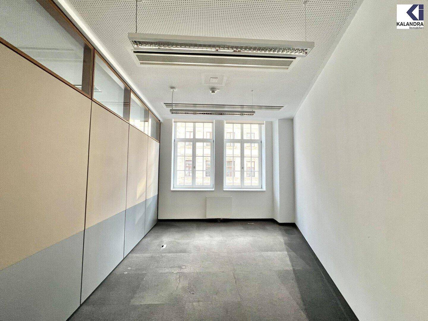 Kleinerer Büroraum mit zwei Fenstern und Teppichboden, angrenzend an eine Trennwand.