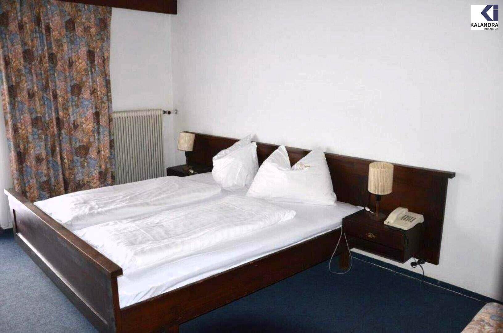 Schlafzimmer mit dunklem Doppelbett, weißen Bettwaren, Nachttischen mit Lampen und Heizkörper an der Wand.