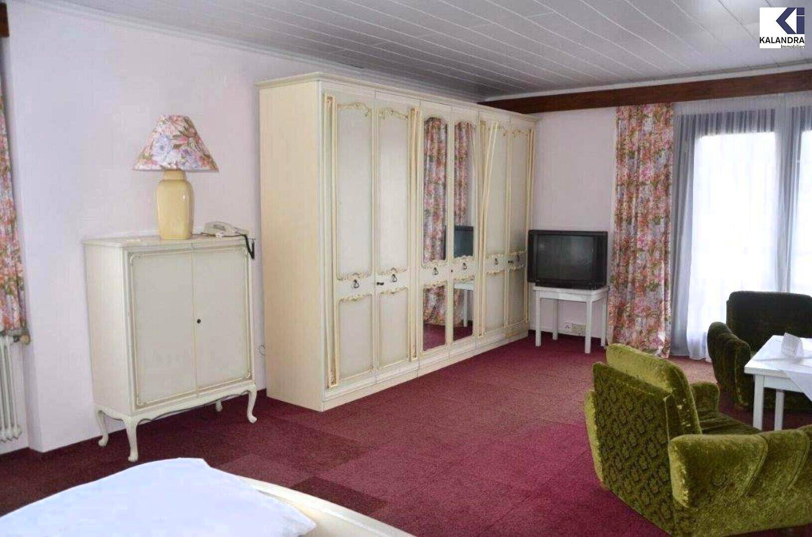 Schlafzimmer mit großem weißen Kleiderschrank, zwei grünen Sesseln, kleinem Tisch und Fernseher auf rotem Teppichboden.