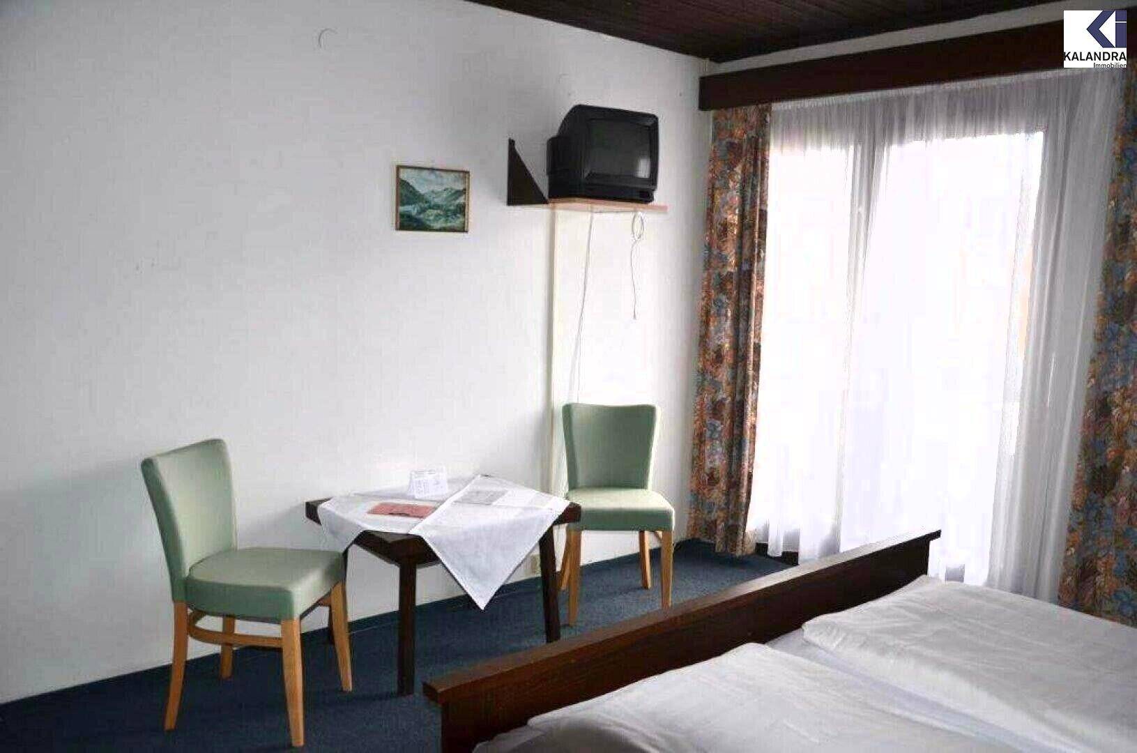 Schlafzimmer mit Doppelbett, zwei grünen Stühlen, kleinem Tisch und älterem Fernseher neben einem großen Fenster.