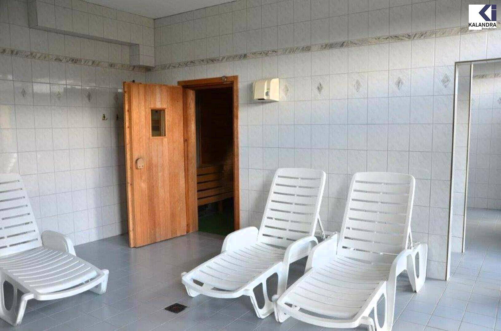 Gefliester Raum mit weißen Liegestühlen und einer Holztür, die zu einer Sauna führt, ideal zum Entspannen.