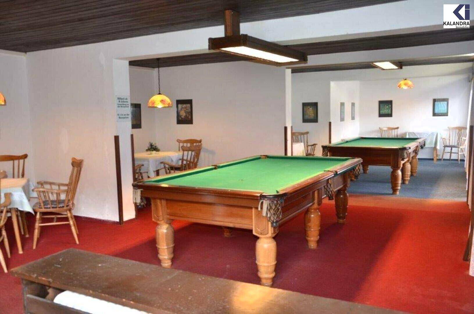 Raum mit zwei Billardtischen, rotem Teppichboden und kleinen Esstischen im Hintergrund unter einer dunklen Holzdecke.