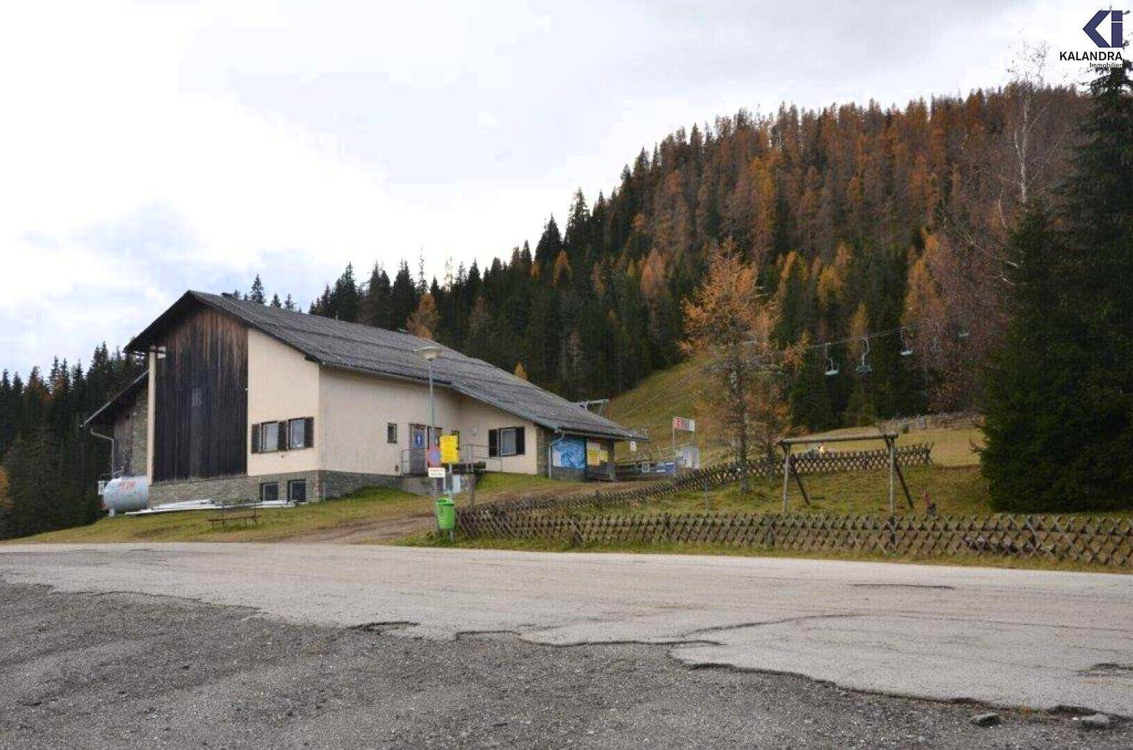 Gebäude in einer bergigen Waldlandschaft mit einem Skilift im Hintergrund unter bewölktem Himmel.