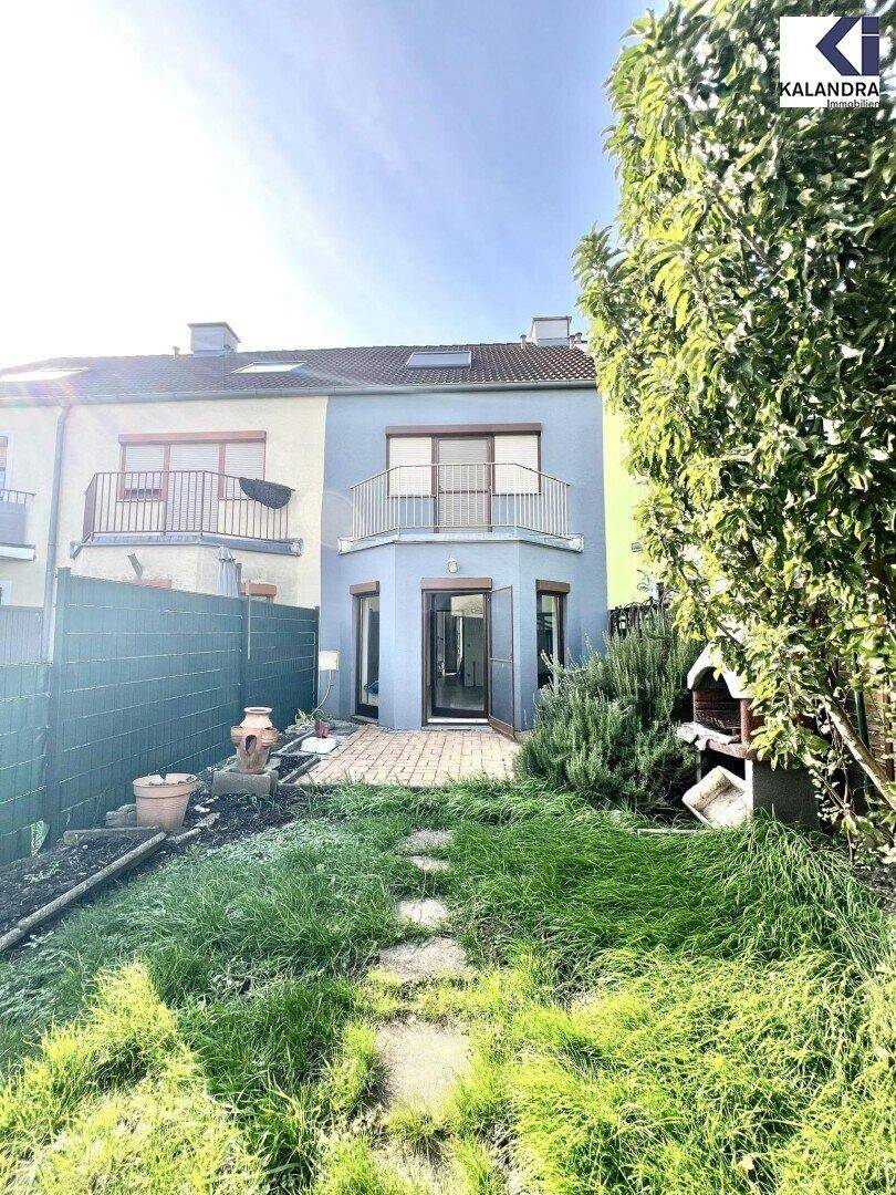 Rückansicht des Reihenhauses mit Garten, Grillplatz und Balkon unter blauem Himmel.