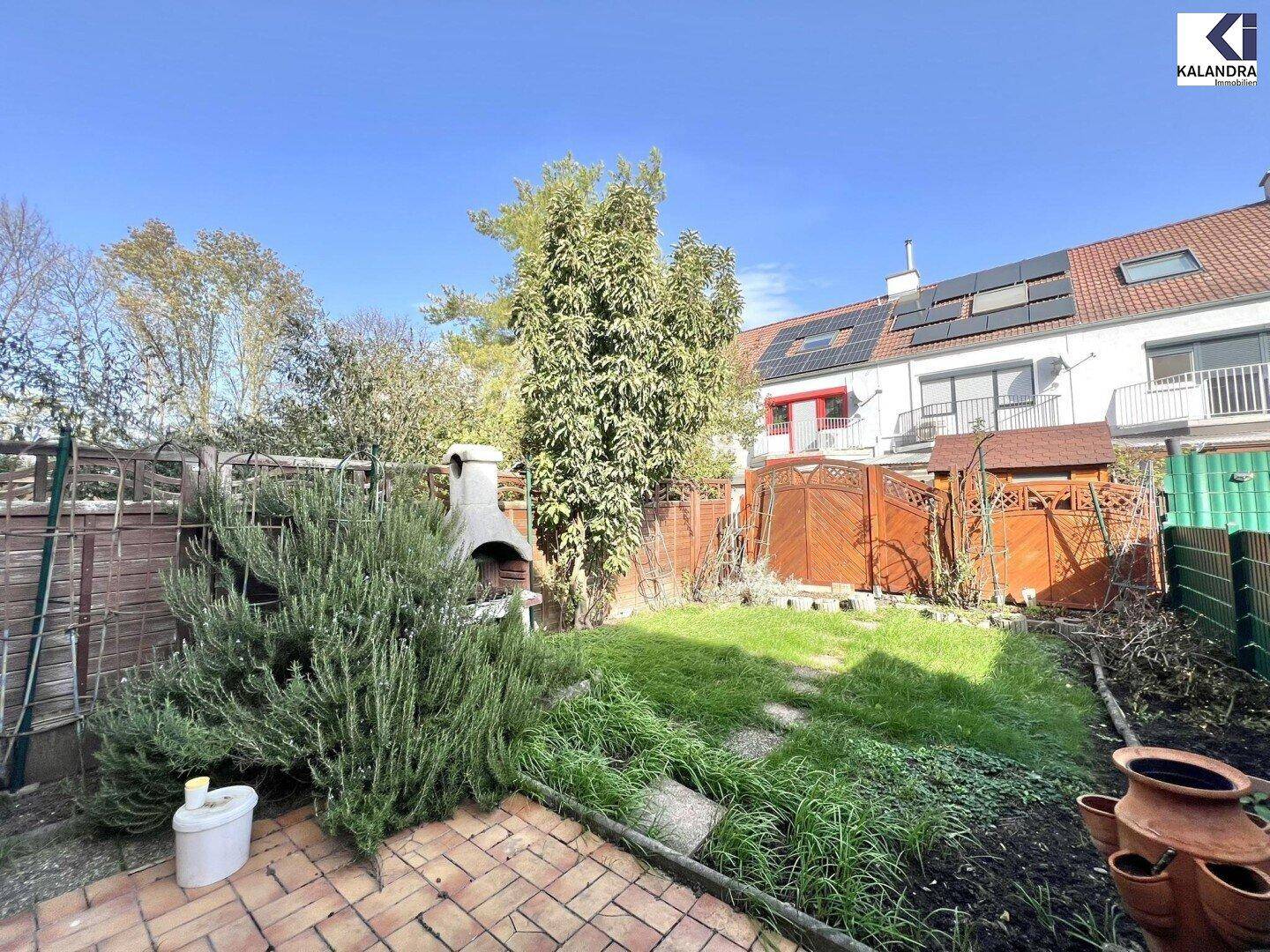 Sonniger Garten mit Grillplatz, Rasen und Blick auf die Rückseite der Reihenhäuser.