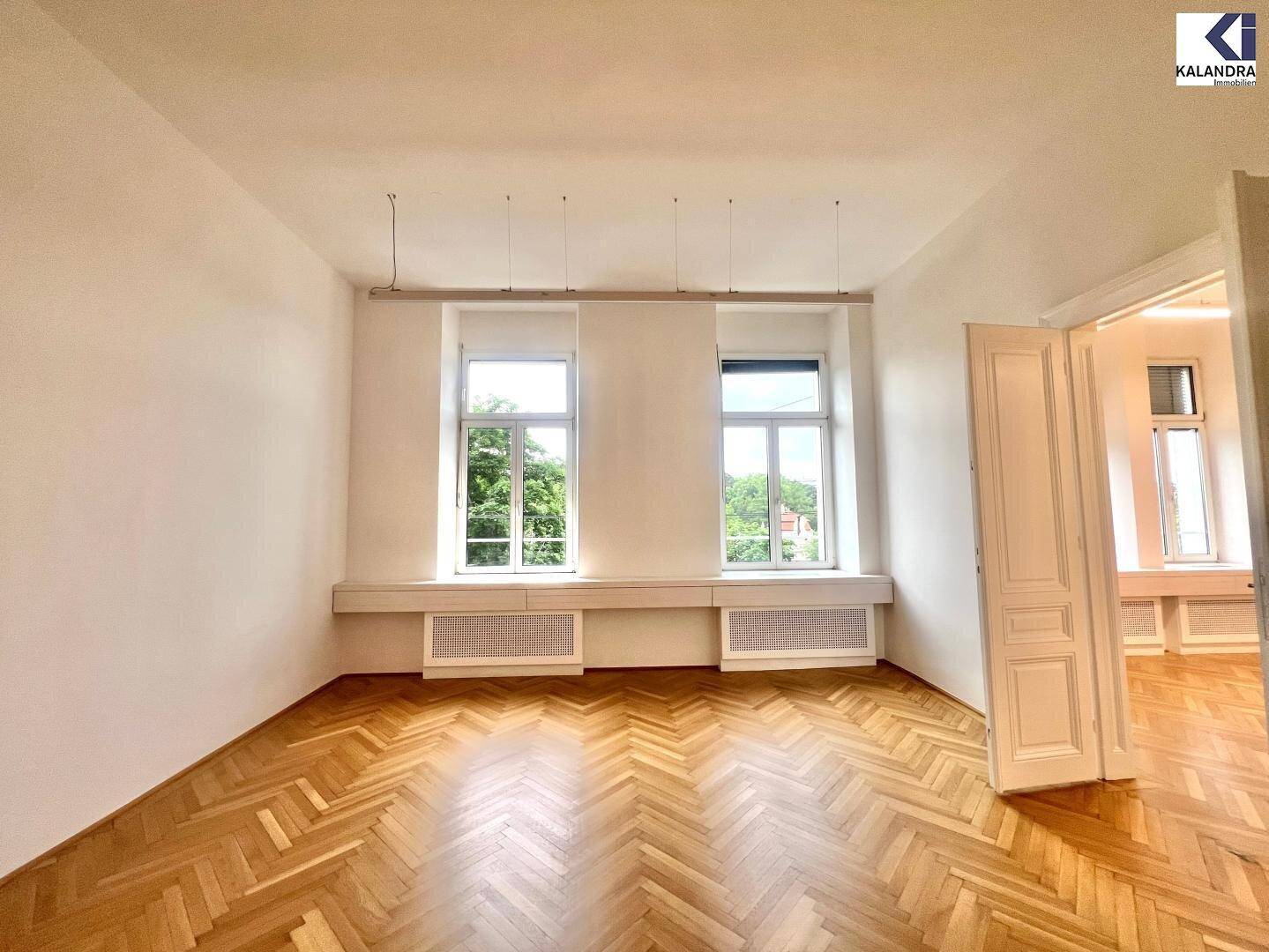 Heller Büroraum mit Fischgrätparkett und zwei hohen Fenstern, die viel Tageslicht hereinlassen.