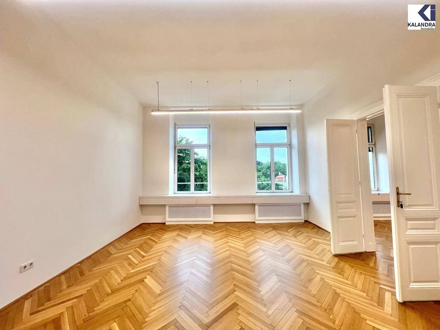Geräumiger Büroraum mit Fischgrätparkett, hohen Fenstern und Blick auf grüne Bäume.