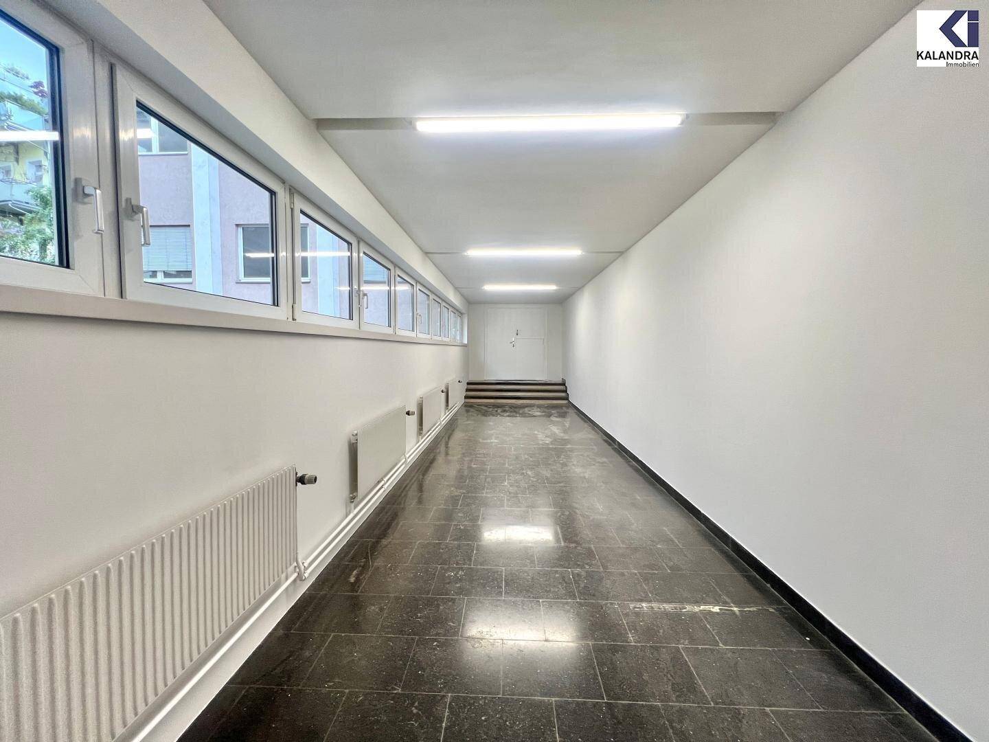 Langer Flur mit dunklem Fliesenboden und Fenstern, die Tageslicht hereinlassen.