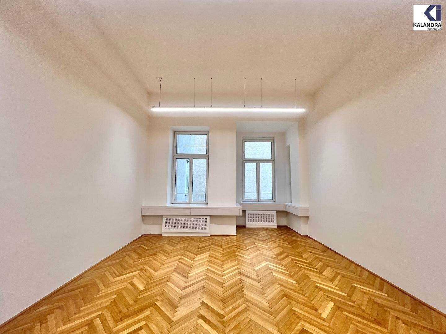 Heller Büroraum mit Fischgrätparkett und zwei hohen Fenstern, die viel Tageslicht hereinlassen.