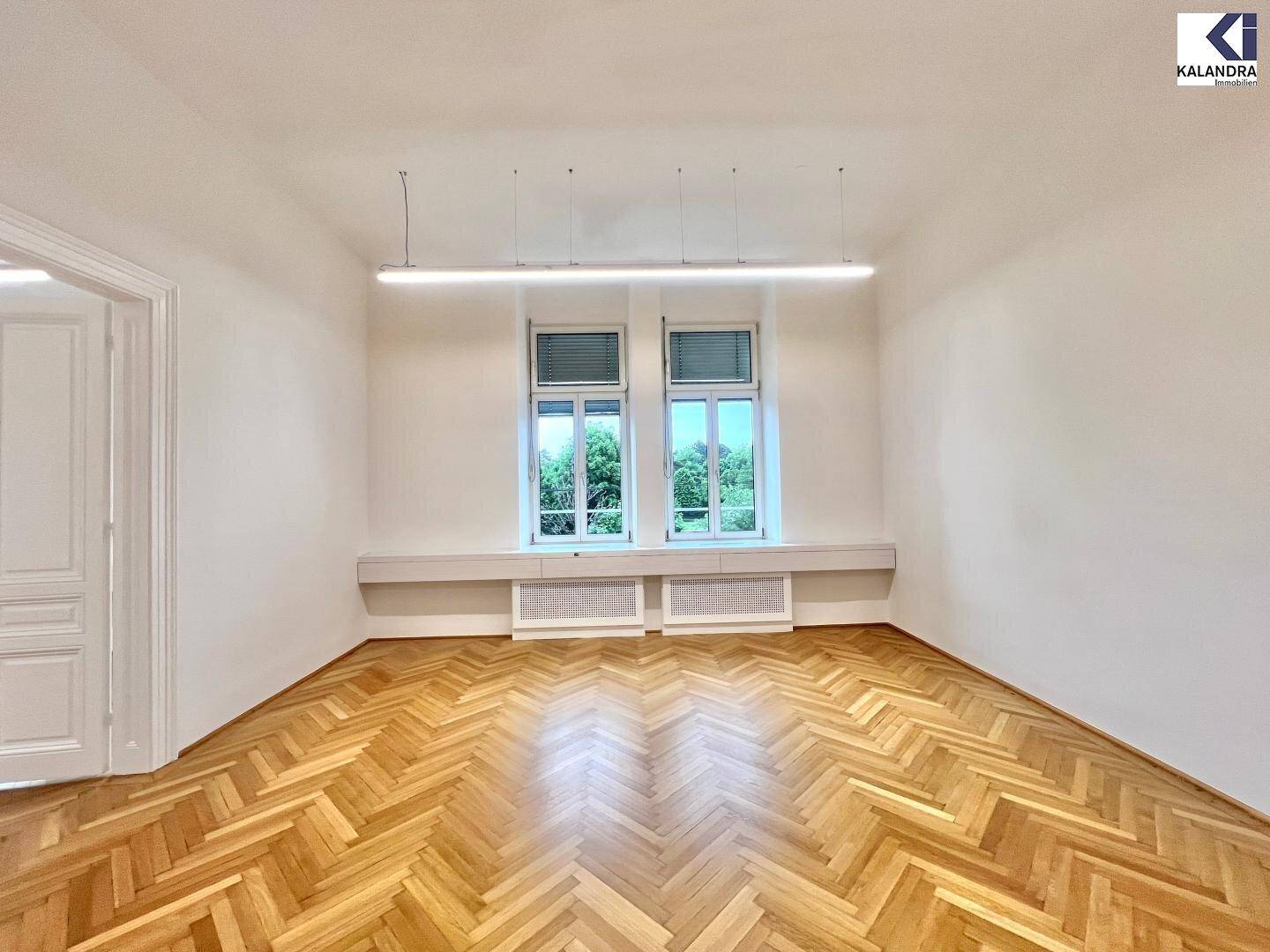 Geräumiger Büroraum mit Fischgrätparkett, hohen Fenstern und Blick auf grüne Bäume.