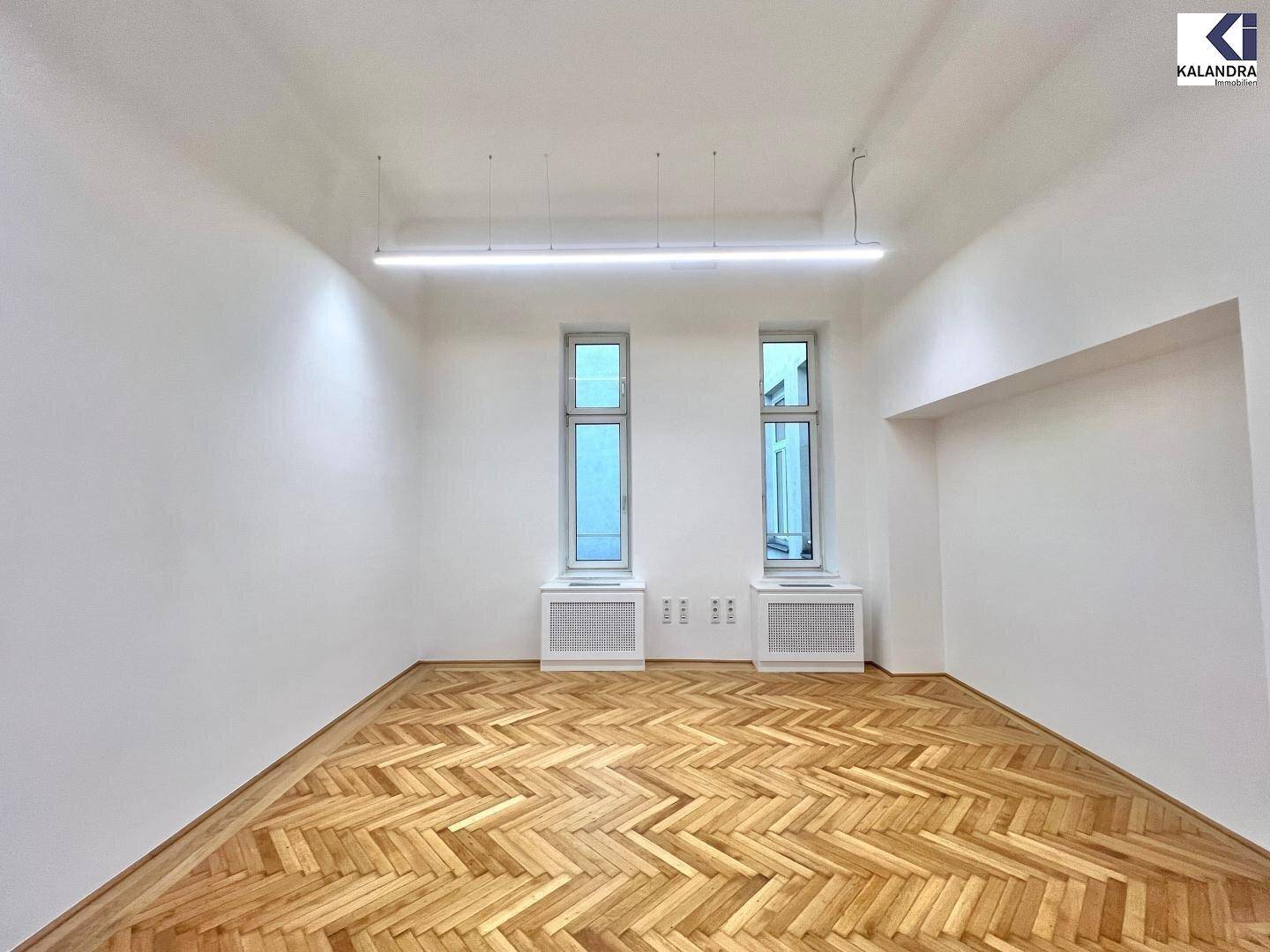Heller Büroraum mit Fischgrätparkett und zwei hohen Fenstern, die viel Tageslicht hereinlassen.