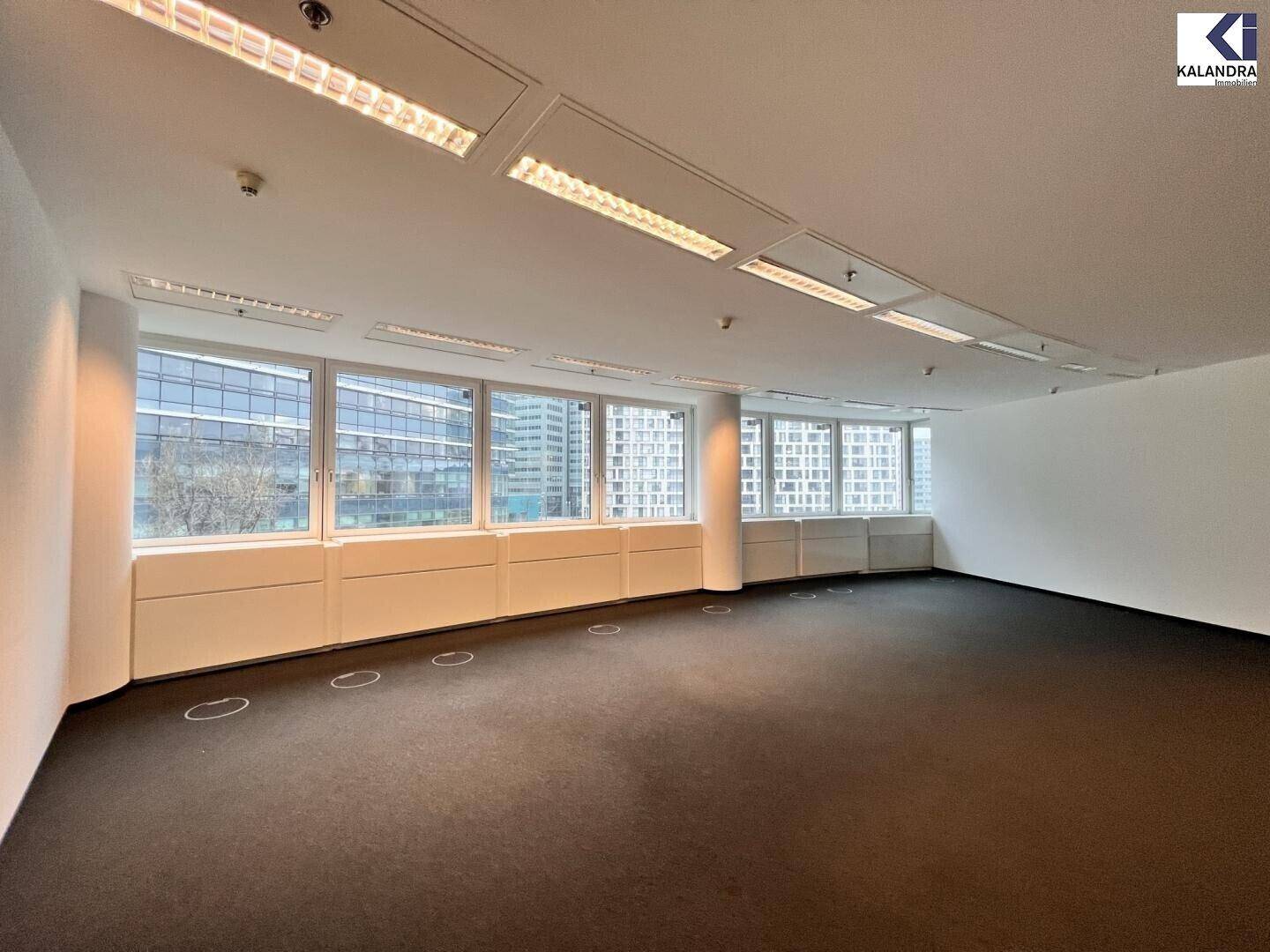 Helles Büro mit durchgehenden Fenstern, Teppichboden und moderner Deckenbeleuchtung in einem Geschäftshaus.