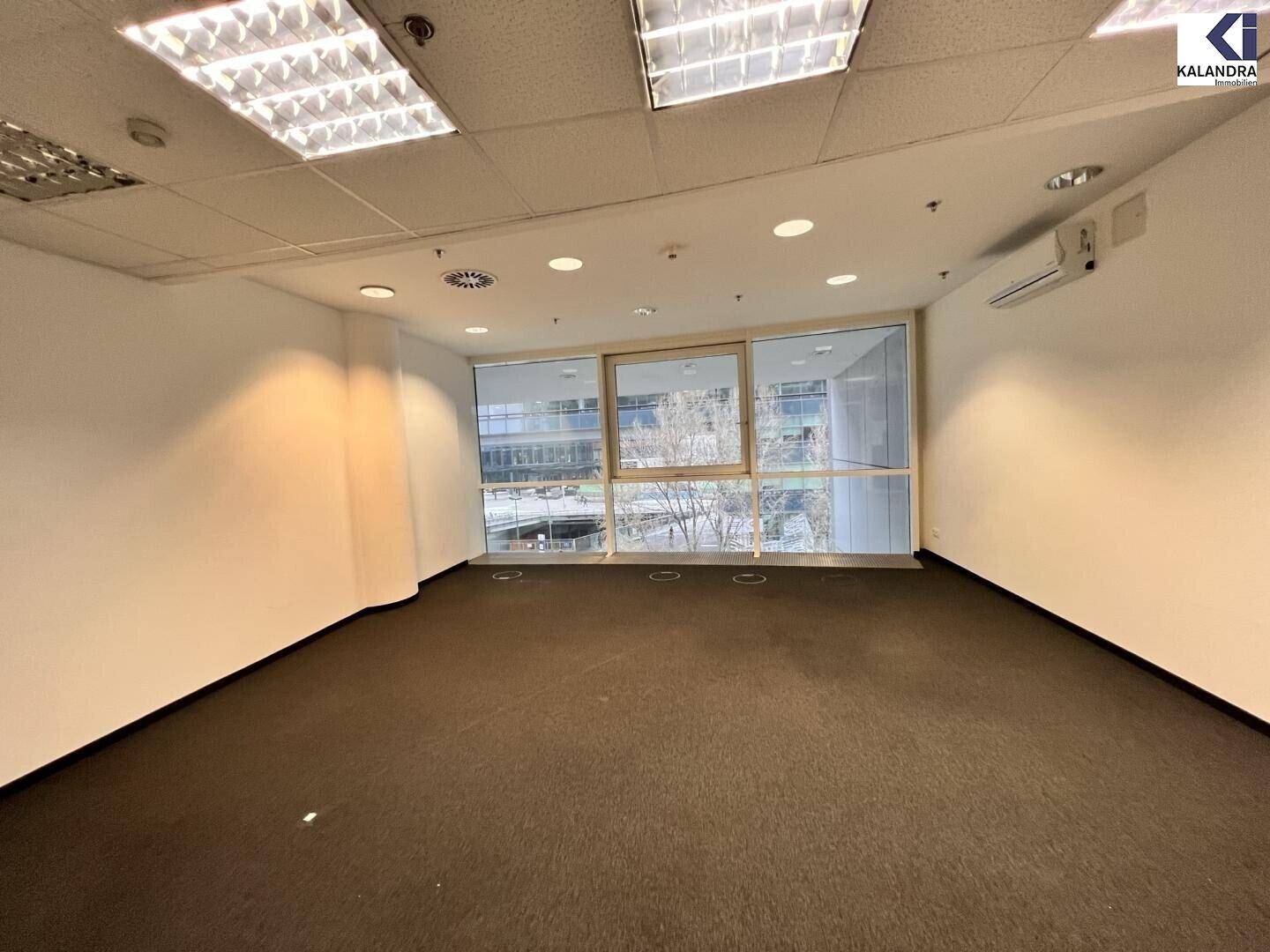 Leeres Büro mit Teppichboden, breiten Fenstern und einer Klimaanlage an der Wand für angenehmes Klima.