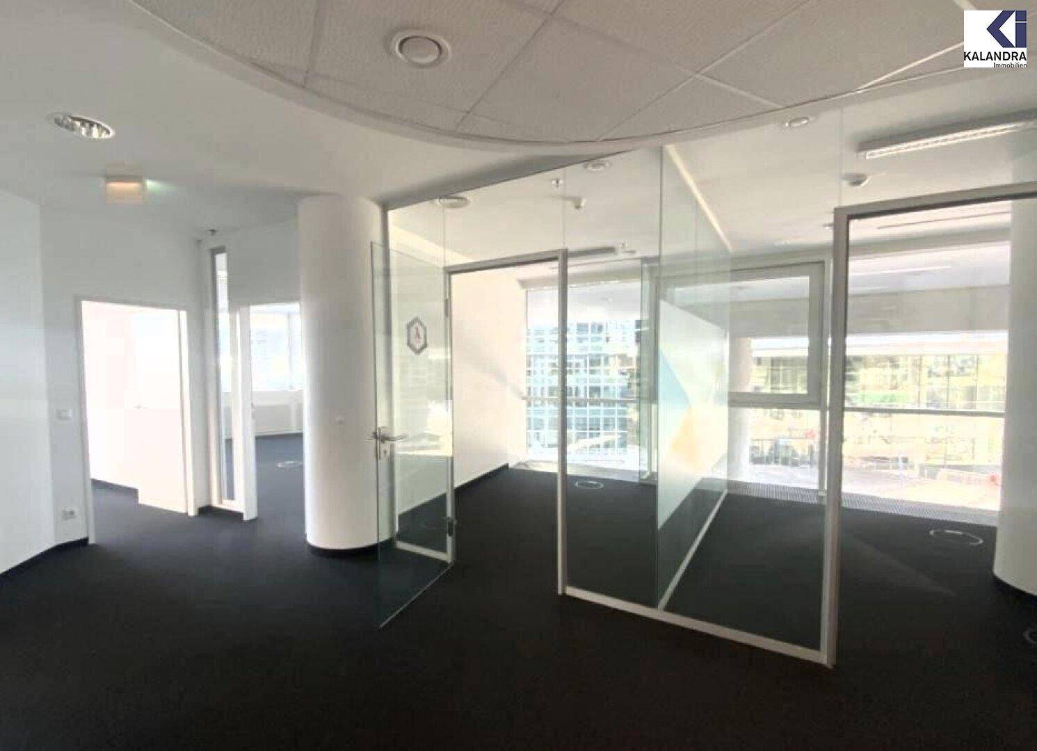 Modernes Büro mit Glaswänden, Teppichboden und viel Tageslicht durch große Fenster.