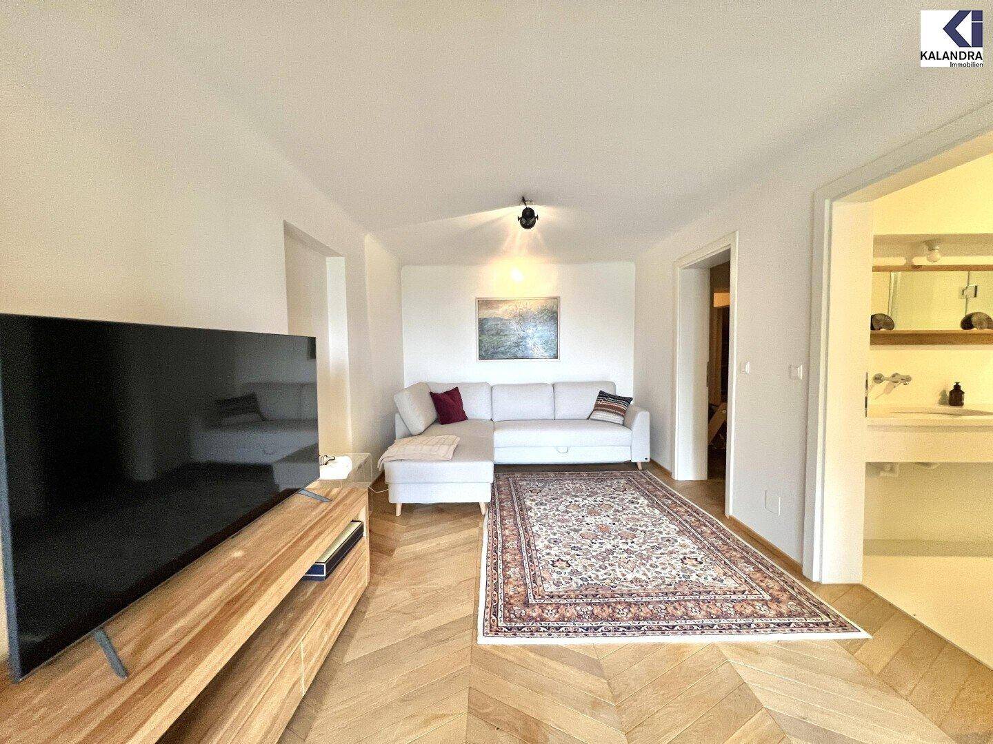 Gemütliches Wohnzimmer mit Sofa, Fernseher und Fischgrätparkettboden, offen zu einem weiteren Bereich.