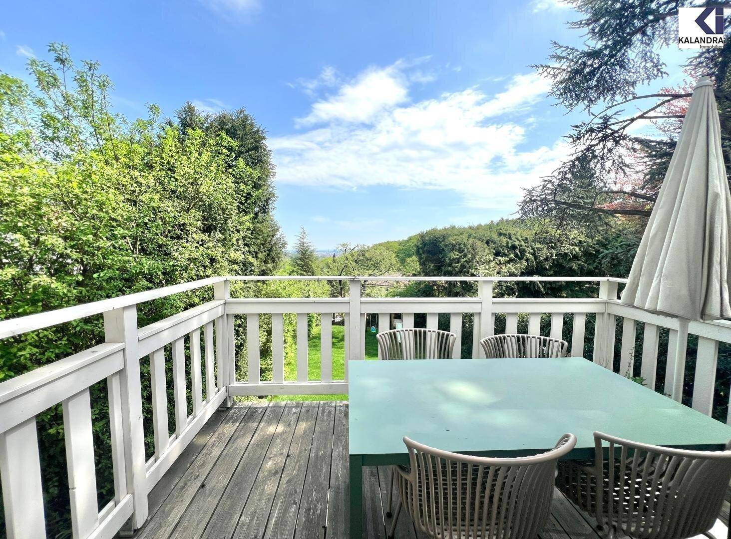 Sonniger Balkon mit Esstisch und Stühlen, umgeben von viel Grün und weitem Ausblick.