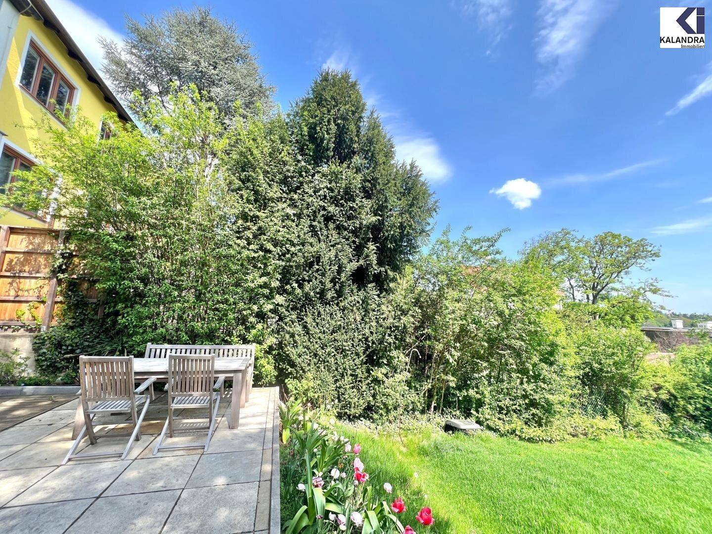 Geräumige Terrasse mit Holztisch und Gartenblick, umgeben von üppigem Grün und Rasenfläche.
