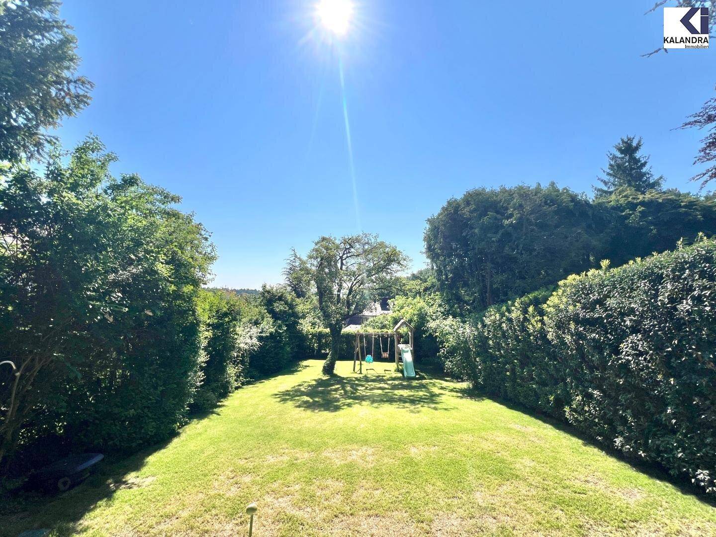 Sonnige Gartenanlage mit weitläufiger Rasenfläche, hohen Hecken und einem Kinderspielplatz.