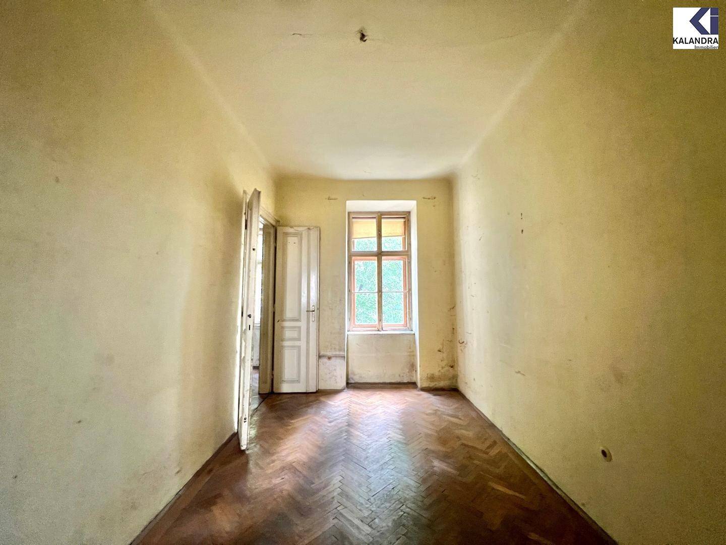 Leerer Raum in sanierungsbedürftigem Zustand mit altem Fischgrätparkett und gelben Wänden.