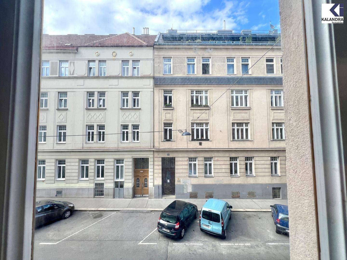 Blick aus dem Fenster auf eine ruhige Stadtstraße mit parkenden Autos und gegenüberliegenden Gebäuden.